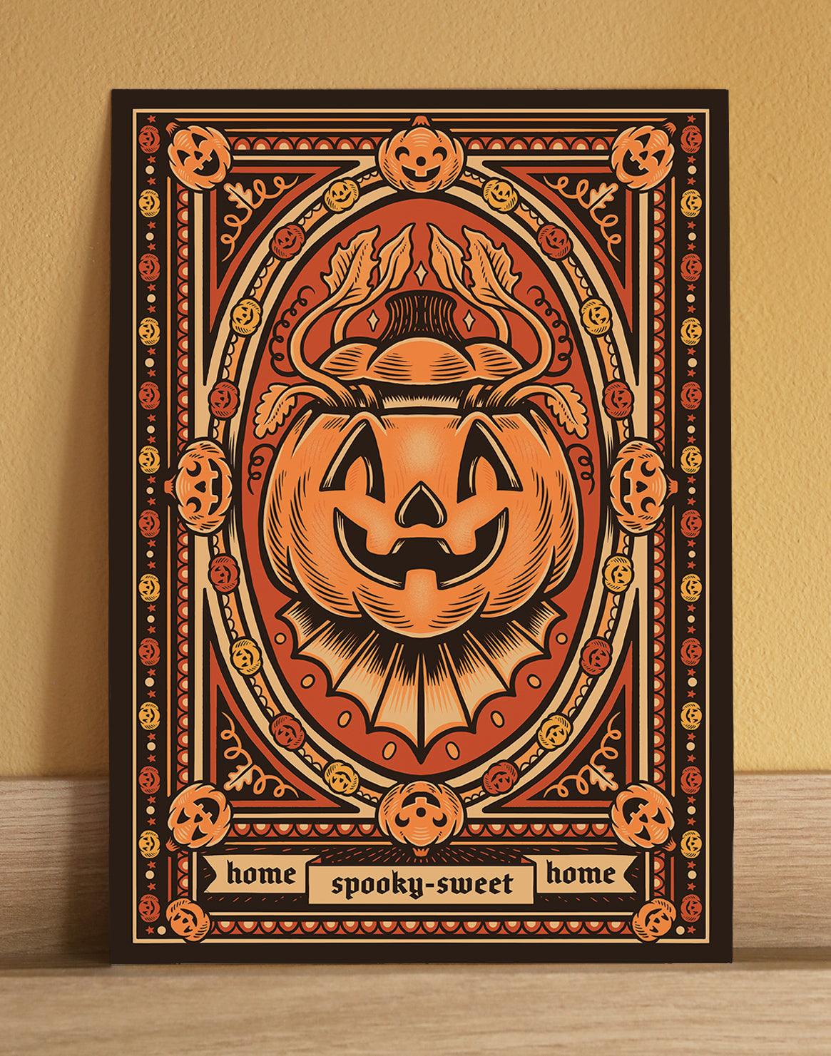 A framed Halloween art print featuring a large carved jack-o'-lantern in the center, surrounded by smaller pumpkins and decorative border motifs. The print has a black frame and is leaning against a beige wall on a light wood surface.