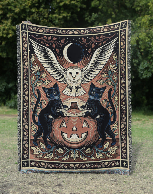 a Halloween tapestry featuring a pumpkin, surrounded by two black cats and a barn owl hovering in the sky below a crescent moon. The tapestry has a floral border. 