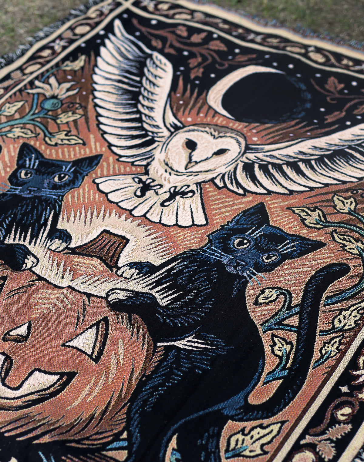 A close up of a halloween tapestry, featuring a jack-o-lantern, black cats and a barn owl with a crescent moon in the sky.