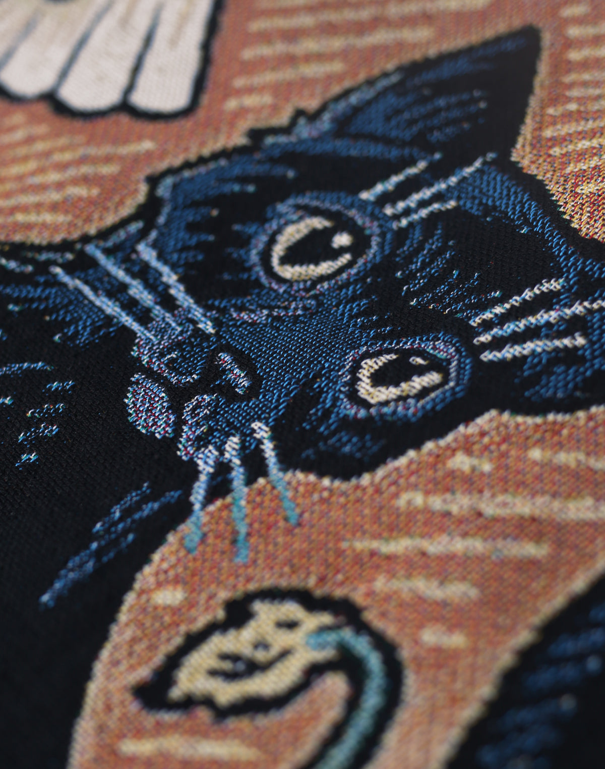 Close-up of a woven textile featuring a black cat face with cream eyes and white whiskers, set against an autumnal tonal background.