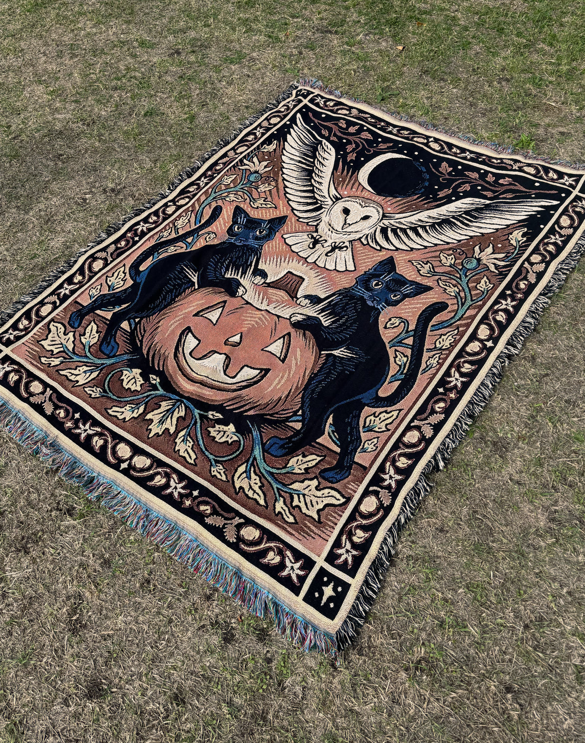 A rectangular woven tapestry with fringe, laid flat on dry grass. The design features a carved orange jack-o-lantern surrounded by two black cats, a barn owl under a crescent moon, and floral motifs in beige, black, blue, and brown.